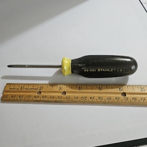 May include: A black and yellow Stanley screwdriver with a flathead tip. The handle is dark with the text "65-301 STANLEY U.S.A." The screwdriver is next to a wooden ruler.