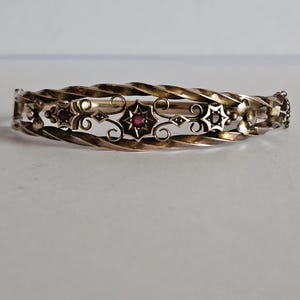 May include: A hinged bangle bracelet featuring a twisted metal band and an openwork center section. The design includes decorative star-shaped motifs, some set with small dark red stones, connected by delicate metal scrollwork and floral accents.