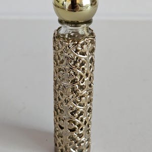 May include: A vintage perfume bottle with a gold-colored spherical cap. The clear glass bottle is encased in an ornate, openwork metal filigree design. The bottle is cylindrical and stands upright.