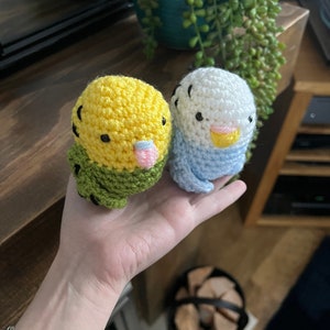 Könnte beinhalten: Zwei gehäkelte Vogel-Plüschtiere, eines gelb und grün, das andere blau und weiß, mit schwarzen Augen und rosa Schnäbeln. Die Plüschtiere werden in einer Hand gehalten.