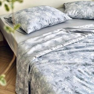 Peut inclure: Une parure de lit compos&eacute;e d'une housse de couette gris clair et de deux taies d'oreiller assorties, orn&eacute;es d'un imprim&eacute; floral botanique blanc. Le tissu pr&eacute;sente un &eacute;clat satin&eacute; lisse, dispos&eacute; sur un lit avec un drap gris uni.