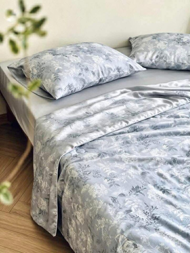 Peut inclure: Une parure de lit compos&eacute;e d'une housse de couette gris clair et de deux taies d'oreiller assorties, orn&eacute;es d'un imprim&eacute; floral botanique blanc. Le tissu pr&eacute;sente un &eacute;clat satin&eacute; lisse, dispos&eacute; sur un lit avec un drap gris uni.