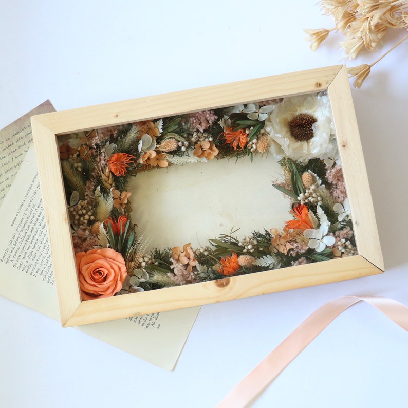 May include: A wooden frame with a dried flower arrangement inside. The flowers are mostly orange and white, with some green foliage. The frame is sitting on a white surface.