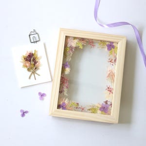 May include: A wooden frame with a pressed flower arrangement inside. The flowers are in shades of pink, purple, and white. The frame is sitting on a white surface.