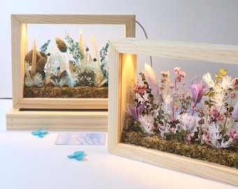 May include: Two rectangular wooden framed art pieces with dried flowers and plants inside. The frames have built-in lights. One frame is on a wooden base. The flowers are in shades of white, green, pink, and purple.