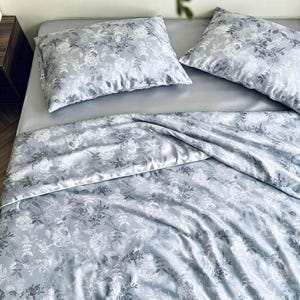 Peut inclure: Un ensemble de literie comprenant deux oreillers rectangulaires et une grande couette de couleur gris clair. Le tissu pr&eacute;sente un imprim&eacute; botanique d&eacute;taill&eacute; avec des motifs floraux et v&eacute;g&eacute;taux blancs et gris fonc&eacute; sur toute la surface.