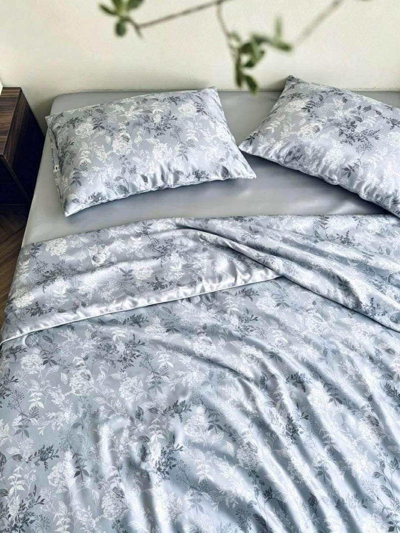 Peut inclure: Un ensemble de literie comprenant deux oreillers rectangulaires et une grande couette de couleur gris clair. Le tissu pr&eacute;sente un imprim&eacute; botanique d&eacute;taill&eacute; avec des motifs floraux et v&eacute;g&eacute;taux blancs et gris fonc&eacute; sur toute la surface.