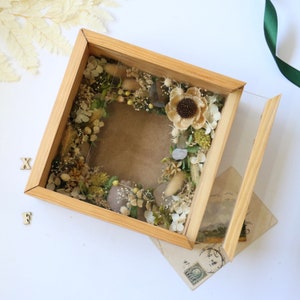 May include: A wooden shadow box with a glass front, containing a dried flower arrangement. The flowers are mostly white and beige, with some green accents. The box is open, revealing the arrangement inside.