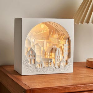 May include: White wooden box with a 3D paper cut-out design of a winter village scene. The scene includes houses, trees, and a moon with stars. The box is illuminated from within, creating a warm glow.