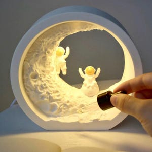May include: White ceramic moon lamp with two astronaut figurines inside. The lamp is lit and has a warm glow. A small bottle of essential oil is being added to the lamp.