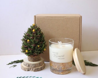 May include: A small artificial pine tree with yellow ornaments sits on a wooden base. Next to it is a glass candle with a white label that reads "Enjoy Every Moment Here & Now". The candle is surrounded by gold glitter.
