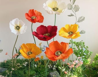 May include: A display of large, colorful paper poppies in shades of white, orange, red, and yellow. The flowers have green stems and are arranged among greenery and small rocks, creating a vibrant floral arrangement.