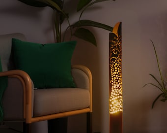 May include: A tall, brown bamboo floor lamp with a decorative cut-out design. The lamp is lit and casting a warm glow. The lamp is in a living room setting with a couch and plants.