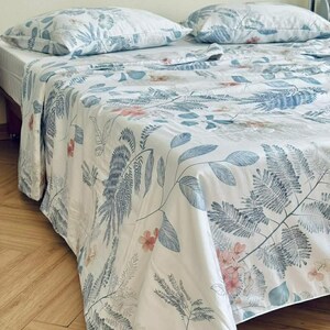 Peut inclure: Un drap de lit blanc et deux oreillers assortis avec un imprim&eacute; botanique de feuilles de foug&egrave;re bleues et de petites fleurs roses. La parure de lit est drap&eacute;e sur un lit, pr&eacute;sentant un motif r&eacute;p&eacute;titif de feuillage et d'&eacute;l&eacute;ments floraux.