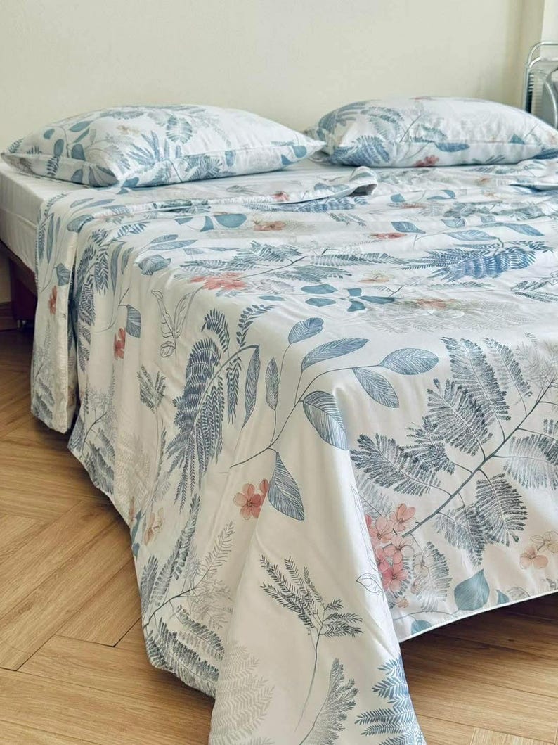 Peut inclure: Un drap de lit blanc et deux oreillers assortis avec un imprim&eacute; botanique de feuilles de foug&egrave;re bleues et de petites fleurs roses. La parure de lit est drap&eacute;e sur un lit, pr&eacute;sentant un motif r&eacute;p&eacute;titif de feuillage et d'&eacute;l&eacute;ments floraux.
