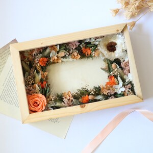 May include: A wooden frame with a dried flower arrangement inside. The arrangement features orange and white flowers, dried grasses, and greenery.