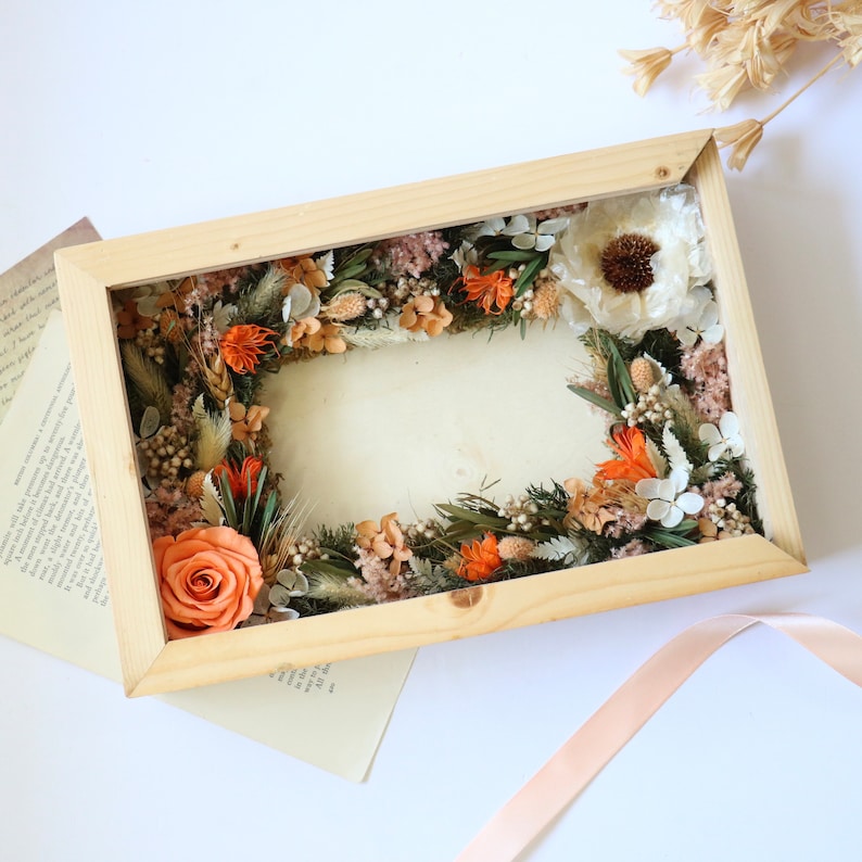 May include: A wooden frame with a dried flower arrangement inside. The arrangement features orange and white flowers, dried grasses, and greenery.