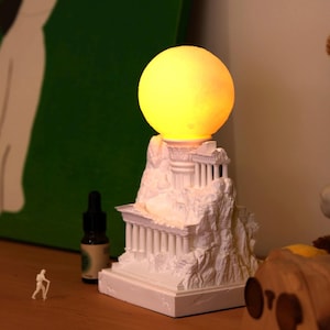 May include: White ceramic lamp shaped like a Greek temple with a glowing yellow moon on top. The lamp is on a wooden surface.