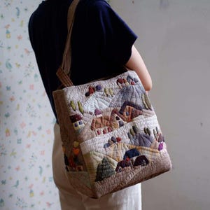 Puede incluir: Un bolso tote acolchado beige con un diseño de paisaje. El bolso presenta un diseño de patchwork con casas, árboles y colinas en varios colores. El bolso tiene una correa marrón y se lleva al hombro.