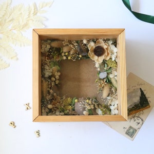 May include: A wooden shadow box with a dried flower arrangement inside. The flowers are a mix of white, brown, and green. The box is sitting on a white surface.