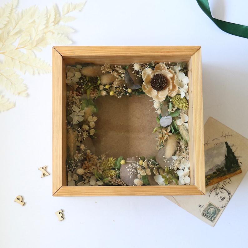 May include: A wooden shadow box with a dried flower arrangement inside. The flowers are a mix of white, brown, and green. The box is sitting on a white surface.