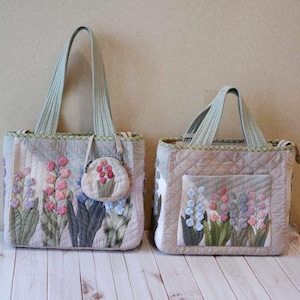 以下が含まれることがあります： 花柄刺繍が施された、2つのキルティングトートバッグ。大きなバッグには長い持ち手があり、小さな丸い小銭入れが付いています。小さなバッグには短い持ち手があります。どちらのバッグも、パステルカラーの花のアップリケが付いたキルティングデザインが特徴です。