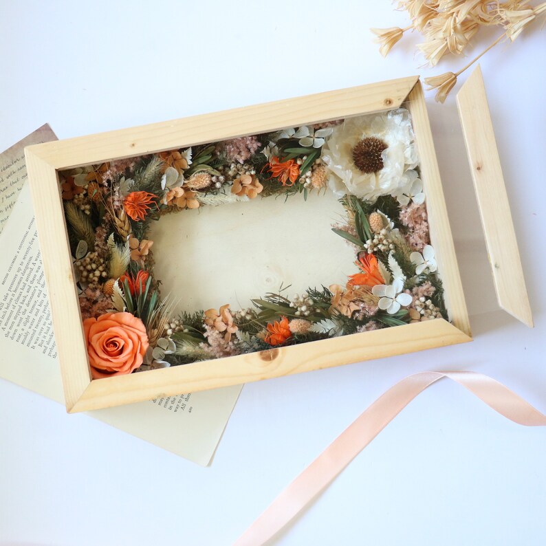 May include: A wooden frame with a glass front containing a dried flower arrangement. The arrangement features a variety of dried flowers in shades of orange, white, and brown. The frame is open on one side, revealing the back of the frame.