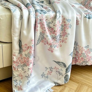 Peut inclure: Un couvre-lit blanc orn&eacute; d'un motif floral r&eacute;p&eacute;titif compos&eacute; de grappes d'hortensias rose tendre et bleu sourd avec des feuilles vertes. Le tissu pr&eacute;sente une finition lisse et l&eacute;g&egrave;rement r&eacute;fl&eacute;chissante, drap&eacute; sur un lit avec un bord passepoil&eacute;.
