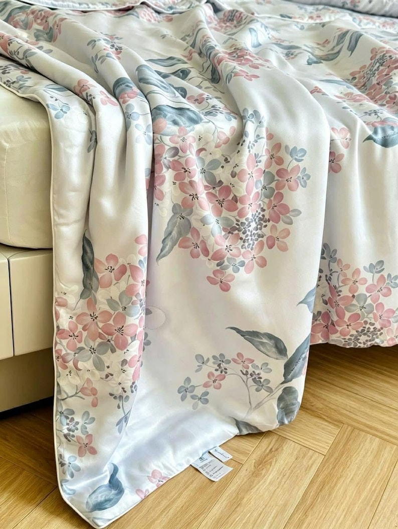 Peut inclure: Un couvre-lit blanc orn&eacute; d'un motif floral r&eacute;p&eacute;titif compos&eacute; de grappes d'hortensias rose tendre et bleu sourd avec des feuilles vertes. Le tissu pr&eacute;sente une finition lisse et l&eacute;g&egrave;rement r&eacute;fl&eacute;chissante, drap&eacute; sur un lit avec un bord passepoil&eacute;.
