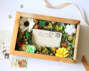 May include: A wooden shadow box filled with dried flowers and greenery, including a white daisy, a green rose, a yellow flower, and a cotton boll. A vintage postcard with the text "Post Card" is centered in the box.