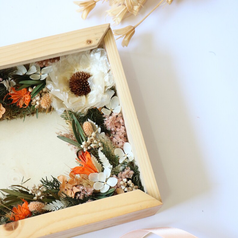 May include: A wooden frame filled with dried flowers in shades of white, orange, and brown. The flowers are arranged in a circular pattern and are surrounded by a thin, wooden frame.