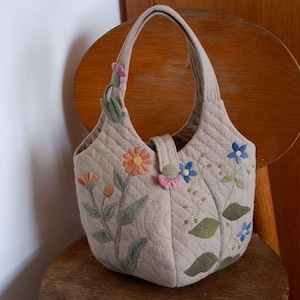 May include: Beige quilted handbag with floral embroidery. The bag has a curved handle and a flap closure. The embroidered flowers are in shades of orange, blue, pink, and green. The bag is on a wooden chair.
