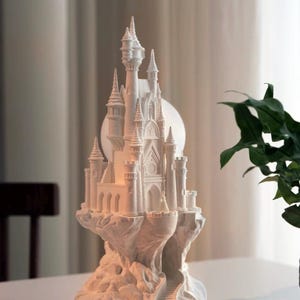 May include: A white ceramic castle figurine with intricate details, perched on a rocky base. The castle has multiple towers and a pointed roof, resembling a fairytale castle.