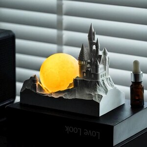 May include: A white 3D printed castle figurine with a glowing yellow moon behind it. The castle is on a rocky base and the moon is on a separate base. The figurine is on a black surface with the text "LOVE LOOK" printed on it.