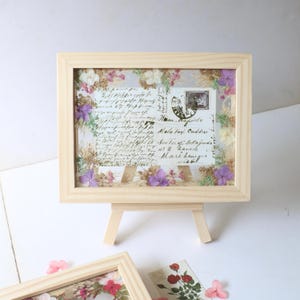 May include: A wooden frame with a vintage postcard inside. The postcard has handwritten text and a stamp. The frame is decorated with pressed flowers in shades of purple, white, and green. The frame is on a small wooden easel.