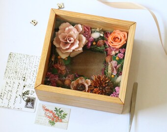 May include: A wooden shadow box with a glass front, containing a dried flower arrangement. The arrangement features a variety of flowers in shades of pink, orange, and brown, including roses, berries, and dried grasses.