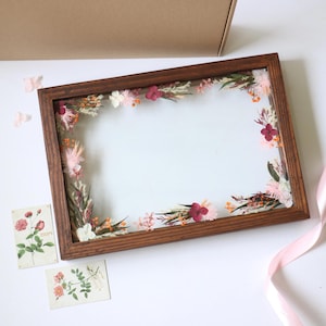 May include: A rectangular wooden floating frame containing pressed dried flowers in shades of pink, burgundy, and orange. Two small rectangular cards featuring botanical illustrations of roses are placed near the frame, alongside a light pink ribbon.