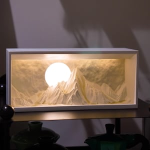 Könnte beinhalten: Eine weiße rechteckige Box mit einer beleuchteten Szene einer Bergkette und einem großen, runden Mond. Die Szene ist aus strukturiertem Papier gefertigt und wird von hinten beleuchtet.