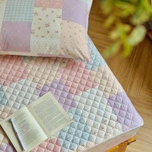 May include: A patchwork quilt and pillow set in pastel colors of pink, lavender, and blue. The quilt features a diamond pattern, and the pillow has a patchwork design with floral and plaid patterns. An open book and pen rest on the quilt.