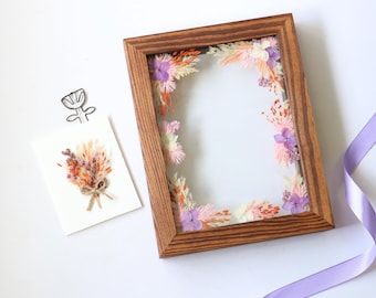 May include: A rectangular wooden frame with a clear glass center, adorned with dried flowers in shades of pink, orange, and purple. A small card with a floral arrangement and a paperclip are next to the frame. A purple ribbon is in the corner.