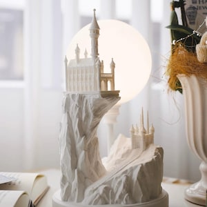 Puede incluir: Escultura de cerámica blanca de un castillo en un acantilado con una luna detrás. El castillo tiene un techo puntiagudo y torres. El acantilado es texturizado y tiene un camino que conduce al castillo.