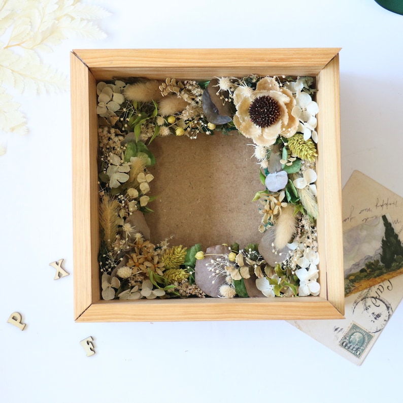 May include: A wooden frame with a dried flower arrangement inside. The flowers are mostly white and beige with some green accents. The frame is sitting on a white surface.