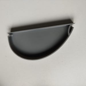 Peut inclure: Un objet gris semi-circulaire imprimé en 3D, avec un dessus plat et un fond incurvé. L'objet a une surface texturée et une petite encoche à une extrémité. Le design est simple et fonctionnel.