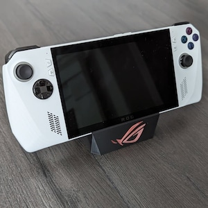 Op de afbeelding: Een witte handheld gameconsole met een zwart scherm, zwarte knoppen en een zwarte joystick. De console staat op een zwarte standaard met het ROG-logo in rood. De console heeft een strak, modern design.