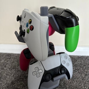 May include: A gray 3D-printed gaming controller stand holding four game controllers. The controllers are white, black, red, and green. The stand is designed to hold Xbox and PlayStation controllers.