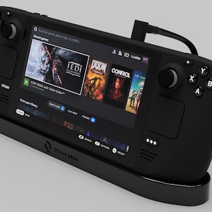 May include: Black Steam Deck handheld gaming console with a screen displaying game titles. The console is docked in a black charging station. The screen shows the Steam logo and the words "STEAM DECK".