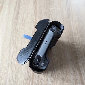 Puede incluir: Una funda protectora negra, abierta para revelar un dispositivo electrónico gris. La funda tiene una superficie texturizada y una pestaña azul. El dispositivo tiene un botón y una luz roja. La funda y el dispositivo están sobre una superficie de madera clara.