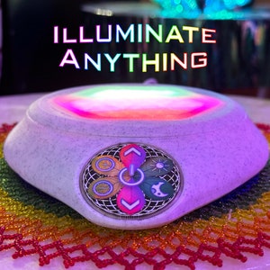 Op de afbeelding: Een witte ronde lichtbasis met een regenboogkleurig licht dat uit het midden straalt. De basis heeft een bedieningspaneel met vijf knoppen, elk met een ander symbool. De tekst "ILLUMINATE ANYTHING" wordt boven de basis weergegeven.
