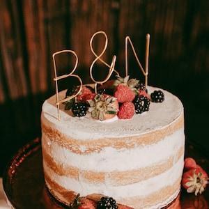 May include: A naked cake with white frosting and a gold wire topper that says "B&N". The cake is decorated with fresh raspberries and blackberries.