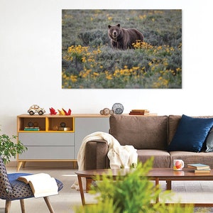 May include: A framed canvas print featuring a brown bear in a field of yellow wildflowers. The bear is centered in the image, surrounded by green foliage and blooms. The artwork is displayed above a modern living room setup.